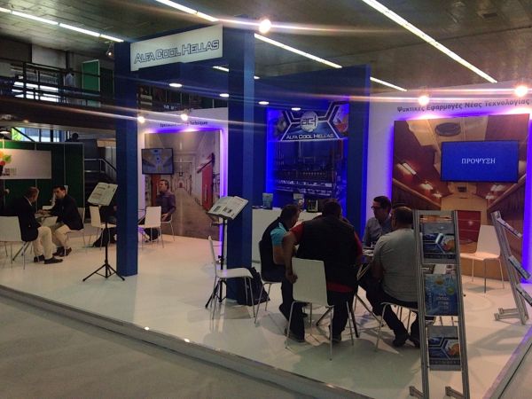 Έκθεση της Alpha Cool Hellas με άτομα στα τραπέζια, διαφημιστικό υλικό και οθόνη που δείχνει 'ΠΡΟΪΟΝΤΑ